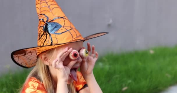 Söt liten kaukasiska flicka med blont hår sju år gammal i dräkt av häxa med hatt och svart orange klänning firar Halloween utomhus på gatan. Semesterkoncept. Långsamma rörelser. — Stockvideo