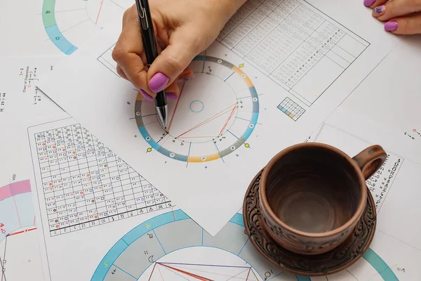 Modern bir astrologun işyeri. Masada duran astrolojik çizelgeler, tablolar, tablolar, diyagramlar. Kadın bir astrologun elleri. Bir fincan çay tabağında.