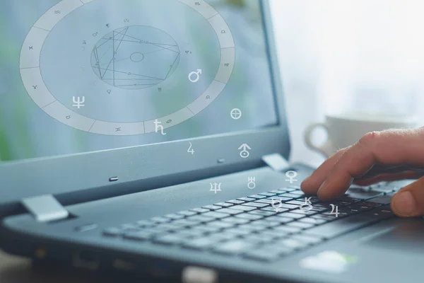 The workplace of a modern advanced astrologer. Laptop on the table for calculating and displaying ephemeris, astrograms and astrological charts. Zodiac circles in the air, astrological signs and symbols of planets and constellations.