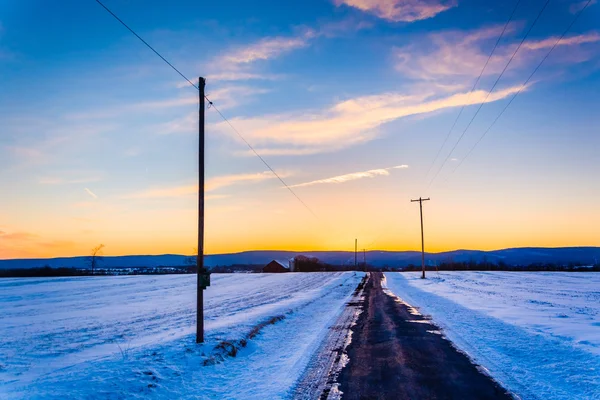 Tramonto su una strada di campagna attraverso campi innevati in campagna — Foto Stock