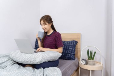 Yatakta dizüstü bilgisayar kullanan Asyalı kadın evde çalışmaktan, uzaktan çalışmaktan ya da evden çalışmaktan keyif alıyor..