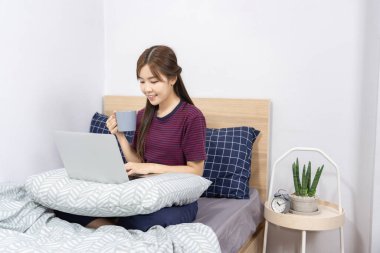 Yatakta dizüstü bilgisayar kullanan Asyalı kadın evde çalışmaktan, uzaktan çalışmaktan ya da evden çalışmaktan keyif alıyor..