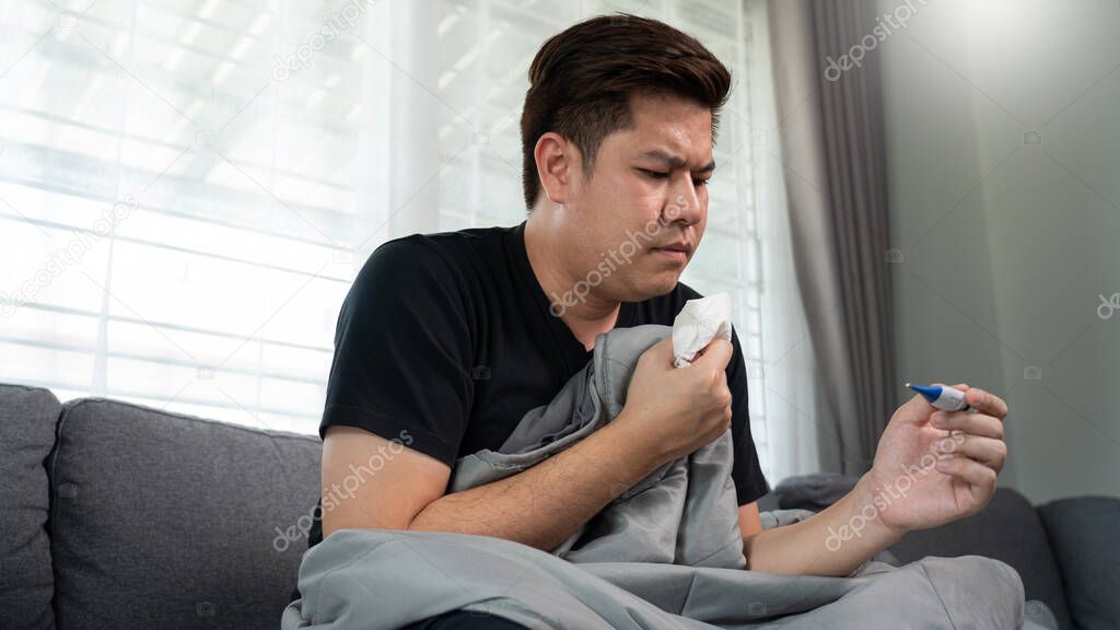 Sick Asian man with flu virus lying in sofa with high fever and measuring body temperature by thermometer and touching his forehead at home