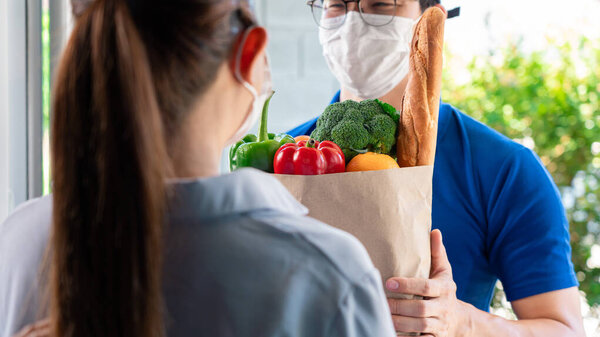 Asian deliver man wearing face mask in uniform handling bag of food and fruit give to woman costumer in front of the house, Express delivery service quarantine and shopping concept