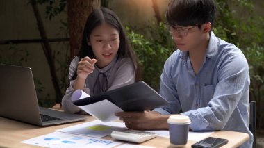 Açık havada çalışan iki erkek ve kadın iş arkadaşı rahatça iş konusunu masaya dizüstü bilgisayar ve belgelerle tartışıyor.