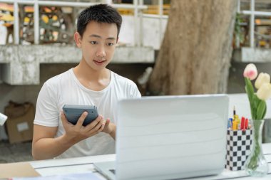 Çevrimiçi konsept üzerinde çalışan beyaz tişörtlü adam oldukça mutlu hissediyor ve hesap makinesini muhasebe sınıfında rakamları hesaplamak için tutuyor..