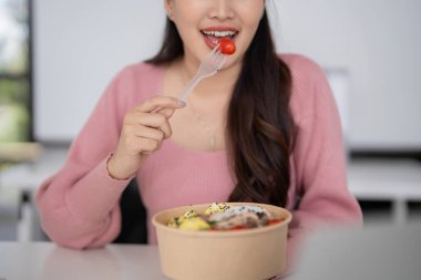 Yetişkin Asyalı kadın taze ve sağlıklı yemeklerden zevk alıyor. Parlak ofis ortamından zevk alıyor. Kişisel refahı profesyonel verimliliği teşvik ediyor.