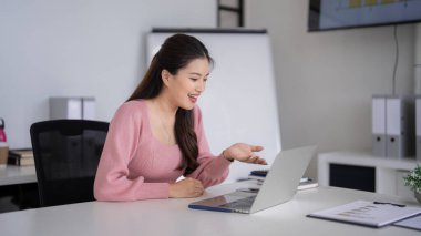 Gülümseyen Asyalı profesyonel kadın online iletişim için dizüstü bilgisayar kullanıyor. Parlak bir ofis ortamında uzaktan çalışma ve sanal buluşma kavramı.