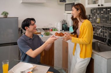 Neşeli Asyalı çift modern mutfakta tatlı çörekleri paylaşıyor. Aile mutluluğu ve şefkat bağı aşikar. Rahat ev hayatı.
