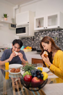 Genç Asyalı yetişkinler kahvaltıyı paylaşır. Mutlu kadın tost yiyor, meyve suyu içiyor. Modern evdeki rahat sabah yaşam tarzı
