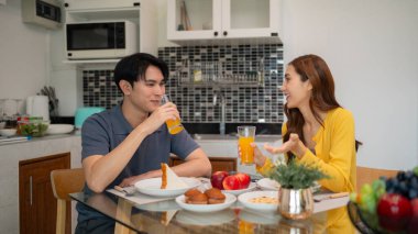 Genç Asyalı yetişkinler evde mutlu bir kahvaltı yaparlar. Meyve suyu hamur işlerini ve sıcacık mutfaklarında sohbet etmeyi seviyorlar. Sabah sıcaklığı birliktelik.