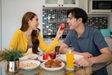 Asyalı ortaklar evde keyifli bir kahvaltı anının tadını çıkarıyorlar. Ona neşe içinde meyve sunuyor. Bağlantı ve refahı arttırıyor.