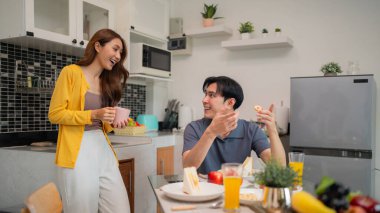 Genç Asyalı çift, parlak çağdaş mutfakta keyifli bir sabah yemeğinin tadını çıkarıyor. Kahkahaları ve rahat bir aile anını paylaşıyorlar..