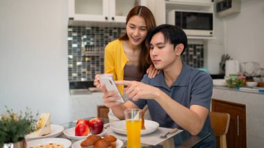 Yetişkin Asyalı ortaklar mobil aygıt tarama içeriğini mutfak masasında paylaşırlar. Günlük yaşam tarzı evde teknolojiyi bağlıyor..