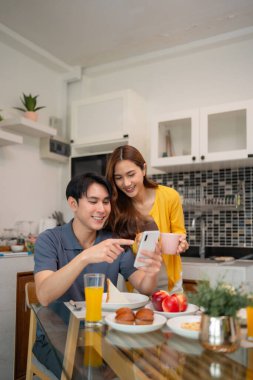 Genç Asyalı yetişkinler ev mutfağında neşeli bir kahvaltı yaparlar. Adam telefonda kadına içerik gösteriyor. Yemek ve içeceklerle rahat bir ev hayatı..
