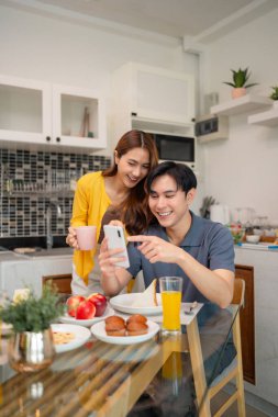 Asyalı yetişkin çiftler evde neşeli bir sabah kahvaltısı paylaşırlar. Dijital içeriğin sosyal etkileşiminden keyif alan bir cep telefonu ekranına bakıyorlar.