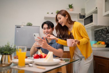 Mutlu Asyalı yetişkin partnerler, neşeli yemek zamanı bağlantı yaşam tarzı sırasında akıllı telefondan ev mutfak masasında dijital içeriğin keyfini çıkarırlar.