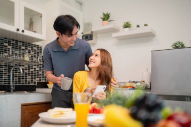 Asyalı çiftler evlerinde samimi bir kahvaltı yaparlar. Modern mutfak ortamlarında akıllı telefon kullanarak gülümserler.