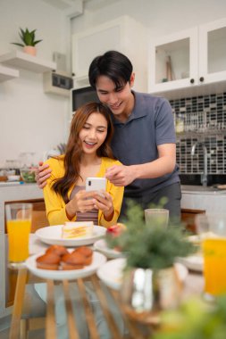 Genç Asyalı yetişkinler telefondaki dijital içeriğin tadını çıkarıyorlar. Samimi evlerinde keyifli bir kahvaltı eşliğinde keyifli anlar paylaşıyorlar..