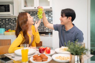 Neşeli Asyalı genç yetişkinler parlak yerli mutfakta sağlıklı kahvaltılık yiyecekler paylaşırlar. Neşeli bir an, taze ürün ile ilişkilerin güçlenmesini sağlar.