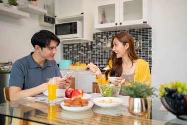 Gülümseyen Asyalı yetişkinler aydınlık bir ev mutfağında rahat bir sabah yemeğini paylaşıyorlar. Taze meyve ve dijital cihazlı sağlıklı yiyecekler sıradan bir evcil ortam yaratır.