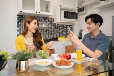 Gülümseyen Asyalı yetişkinler evde besleyici bir yemek ve dijital bağlantının keyfini çıkarıyorlar. Modern mutfak dekorasyonu neşeli bir ev hayatı yansıtır..