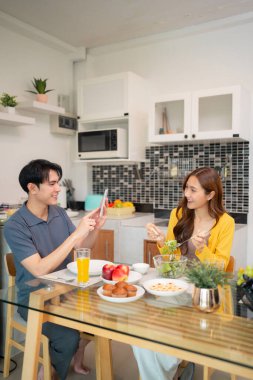Asyalı çift, neşeli bir sabah yemeği fotoğrafı paylaşıyor. Rahat ev sahnesi. Sağlıklı beslenme, pozitif yaşam tarzı bağlantısı.