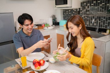 Neşeli Asyalı çift, ev yapımı taze salata hazırlarken mobil cihazlarla uğraşırken ev ortamında mutlu mesut bir yaşam tarzını seviyor.