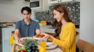 Genç Asyalı yetişkinler, lezzetli çörekler ve meyvelerle sıcak ev ortamları yaratarak ev samimi mutfağında keyifli bir kahvaltı yapıyorlar.