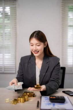 Yetişkin Asyalı kadın, akıllı yatırımı sembolize eden altın külçeleri ve dolar para birimini kendine güvenle elinde tutuyor. Zenginlik yönetimi ve finansal planlama mutlu bir ifadeyle sergilendi.