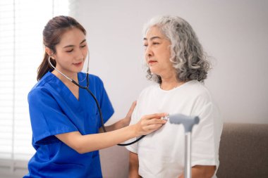 Kendini adamış Asyalı hemşire, daha yaşlı bir hastayı steteskop kullanarak, şefkatli profesyonel sağlık hizmetleri ve destek hizmetleri sunarak özenle muayene ediyor.