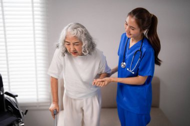 Merhametli Asyalı hemşire, yürüyen yaşlı bir kadına nazikçe yardım eder. Profesyonel bakıcı içeride hareket edebilen hastaları destekliyor.