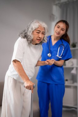 Şefkatli bir bakıcı, bastonuyla yürüyen yaşlı bir Asyalı kadını destekliyor. Ev sağlığı ve hemşirelik hizmetleri gereklidir. Bakıcının yüzü bulanık.
