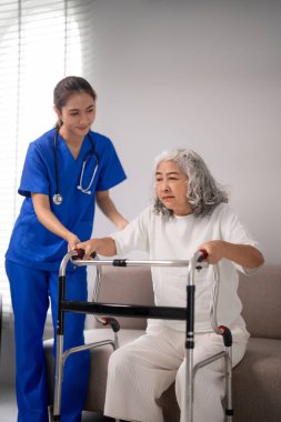 Yetişkin Asyalı bakıcı yaşlı hastaların ayağa kalkmasına Walker 'ı kullanarak yardım ediyor. Yaşlıların günlük yaşam desteği için empati ve profesyonel yardım.