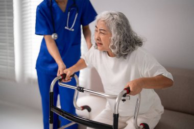 Hemşire yaşlı Asyalı kadın ayakta yürürken nazik bir şekilde yardım eder. Rahat bir ev ortamında bağımsızlığı, saygınlığı ve iyileşmeyi teşvik eder..