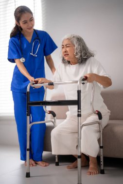 Deneyimli bir Asyalı hemşire, rehabilitasyon sürecinde tıbbi destek için ayakta duran bir yürüteç kullanan yaşlı bir Asyalı kadına dikkatlice yardım eder.