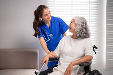 Merhametli Asyalı hemşire yaşlı kadına tekerlekli sandalyede yardım ediyor. Profesyonel bakıcı, evde yaşlılara huzur ve refah sağlamak için nazik destek sunuyor..