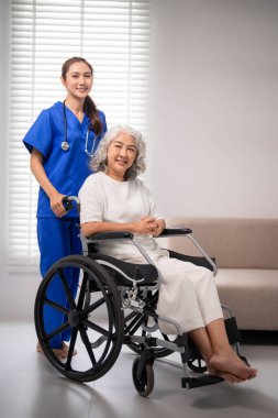 Profesyonel Asyalı hemşire, tekerlekli sandalyedeki yaşlı bir kadın için şefkatli bir bakım sunar. Bu resim, ev sağlığı hizmetleri ve hasta konforunu resmediyor.