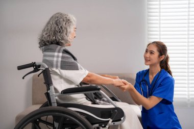 Asyalı hemşire tekerlekli sandalyedeki yaşlı kadına şefkatli bir bakım sunuyor. Profesyonel destek, ev ortamında konfor ve insan ilişkileri sağlar.