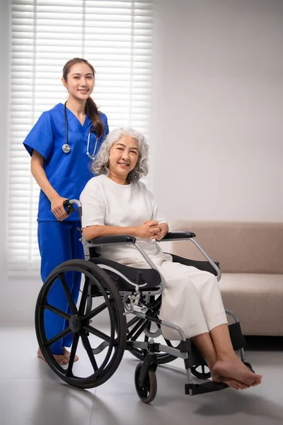 Profesyonel Asyalı hemşire, tekerlekli sandalyedeki yaşlı bir kadın için şefkatli bir bakım sunar. Bu resim, ev sağlığı hizmetleri ve hasta konforunu resmediyor.