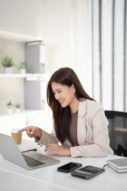 Gülümseyen Asyalı iş kadını, bilgisayardaki verileri hesap makinesi ve akıllı telefonla parlak bir çalışma alanında analiz ediyor. Kişisel finans ya da çevrimiçi işleri yönetir..