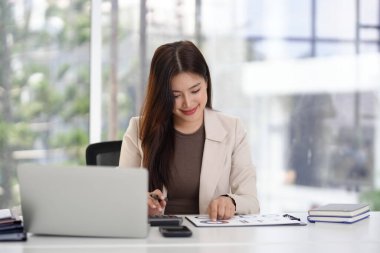 Odaklanmış Asyalı iş kadını finansal bilgi raporlarını analiz ediyor. Verileri hesaplar. Verimli ofis çalışma ortamı için dizüstü bilgisayar kullanır.