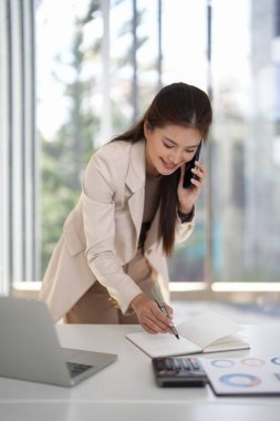 Profesyonel yetişkin Asyalı kadın, iş gününü verimli bir şekilde yönetiyor. Cep telefonu kullanıyor ve modern ofis masasında notlar alıyor..