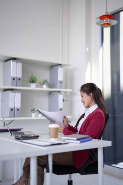 Yetişkin Asyalı kadın yönetici raporları analiz ediyor. Aydınlık ofis alanındaki ergonomik bir masada özenle çalışıyor. Stratejiye odaklanmış.