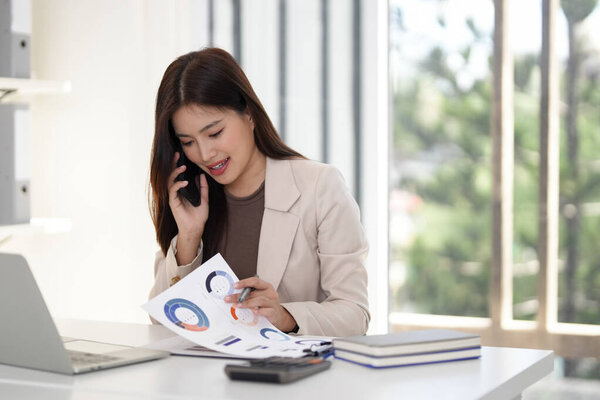 Трудолюбивая азиатская взрослая женщина управляет бизнес-задачами с помощью телефонного звонка, анализирующего рыночные отчеты в современном рабочем пространстве. Эффективная коммуникация способствует повышению производительности.