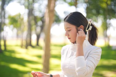 Asyalı kadın, kulaklığı açık hava antrenmanına hazırlıyor. Huzurlu bir parkta sağlık ve zindelik üzerine yoğunlaşırken müziği sever..