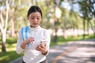 Genç yetişkin Asyalı kadın, sağlık ve aktif yaşam tarzını desteklemek için parkta yapılan egzersizden sonra cep telefonunu kontrol ediyor..