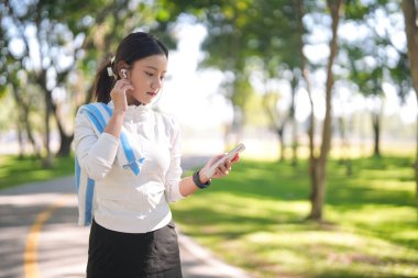 Aktif Asyalı kadın açık hava parkı ayarları kablosuz kulaklıkları düzeltirken akıllı telefonları da spor için gözden geçirir.