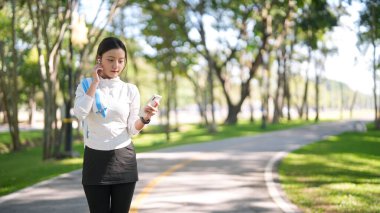 Genç Asyalı yetişkin kadın güzel yeşil parktaki cep telefonunu izlerken kablosuz kulaklığı ayarlıyor. Kişisel fitness ve rahatlama anının tadını çıkarıyorum..
