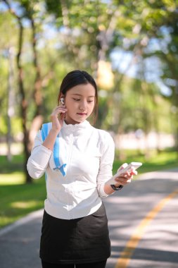 Yetişkin bir Asyalı kadın koşu ya da egzersiz seansına hazırlanıyor. Canlı yeşil park ortamında cep telefonunu kontrol ederken kablosuz kulaklık ayarlıyor..